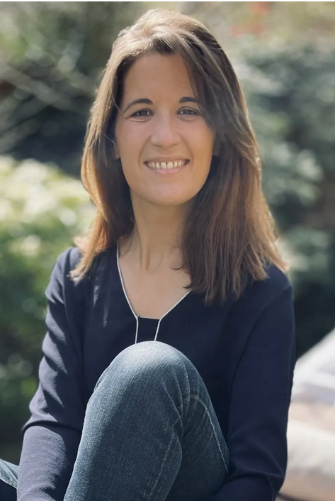 Portrait d’une femme souriante en extérieur, assise en tailleur, vêtue d’un haut bleu foncé et d’un jean