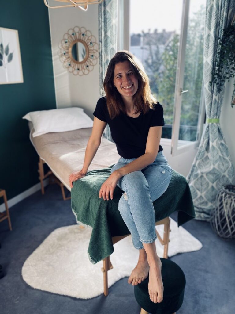 Capucine assise pieds nus sur la table de soin dans son cabinet, souriante, en tenue décontractée